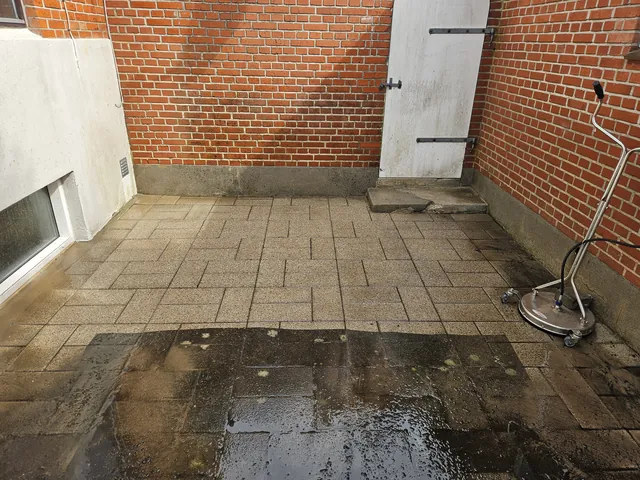 Før fliserens: gårdhave med fliser mellem røde murstensvægge og en hvid dør. Fliserne er mørke af alger og snavs, og en fladerenser til højtryksrenseren står på den våde belægning.
